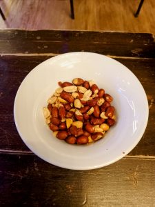 A white ceramic bowl filled with roasted, salted peanuts with their reddish-brown skins partially attached, resting on a dark wooden surface. 