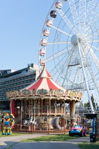 A decorated carousel with a red-and-white striped roof stands in front of a large Ferris wheel in Kraków. Colorful children’s rides and a modern building surround the amusement area under a clear blue sky.
