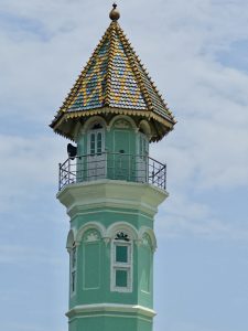 A tall, light-green minaret rises gracefully above the surroundings, featuring arched windows, a balcony, and a striking conical roof decorated with colorful yellow-and-blue patterned tiles. The structure showcases the unique architectural beauty of Ayikkara Muhyidheen Masjid (Moideen Palli). Captured at Kannur, Kerala. 

