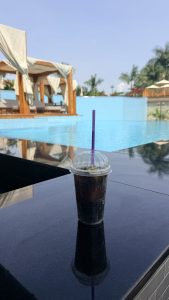 A cold soft drink on a table beside an infinity pool with cabanas and palm trees in the background.