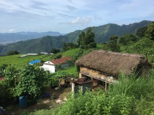 A rustic thatched-roof hut is nestled amidst lush greenery and rolling hills, conveying serenity. Distant mountains and a blue sky complete the pastoral scene.
