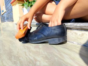 A child polishing a worn black shoe with a wooden brush on a sunlit tiled doorstep. 