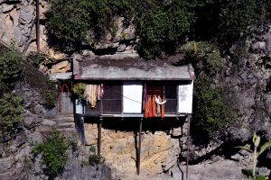 A small home clings to a rocky cliffside, supported by long wooden poles and surrounded by dense greenery. 