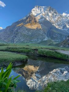 Majestic snow-capped mountain reflecting in a clear stream with lush green foreground under a bright blue sky.