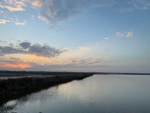 Expansive sunset sky above calm water, with scattered clouds, distant shoreline, and subtle reflections across the surface.
