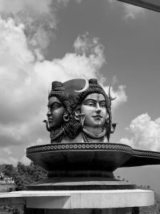 A black and white photograph of a large multi-faced statue of the Hindu deity Lord Shiva with a crescent moon and snakes, set against a cloudy sky.
