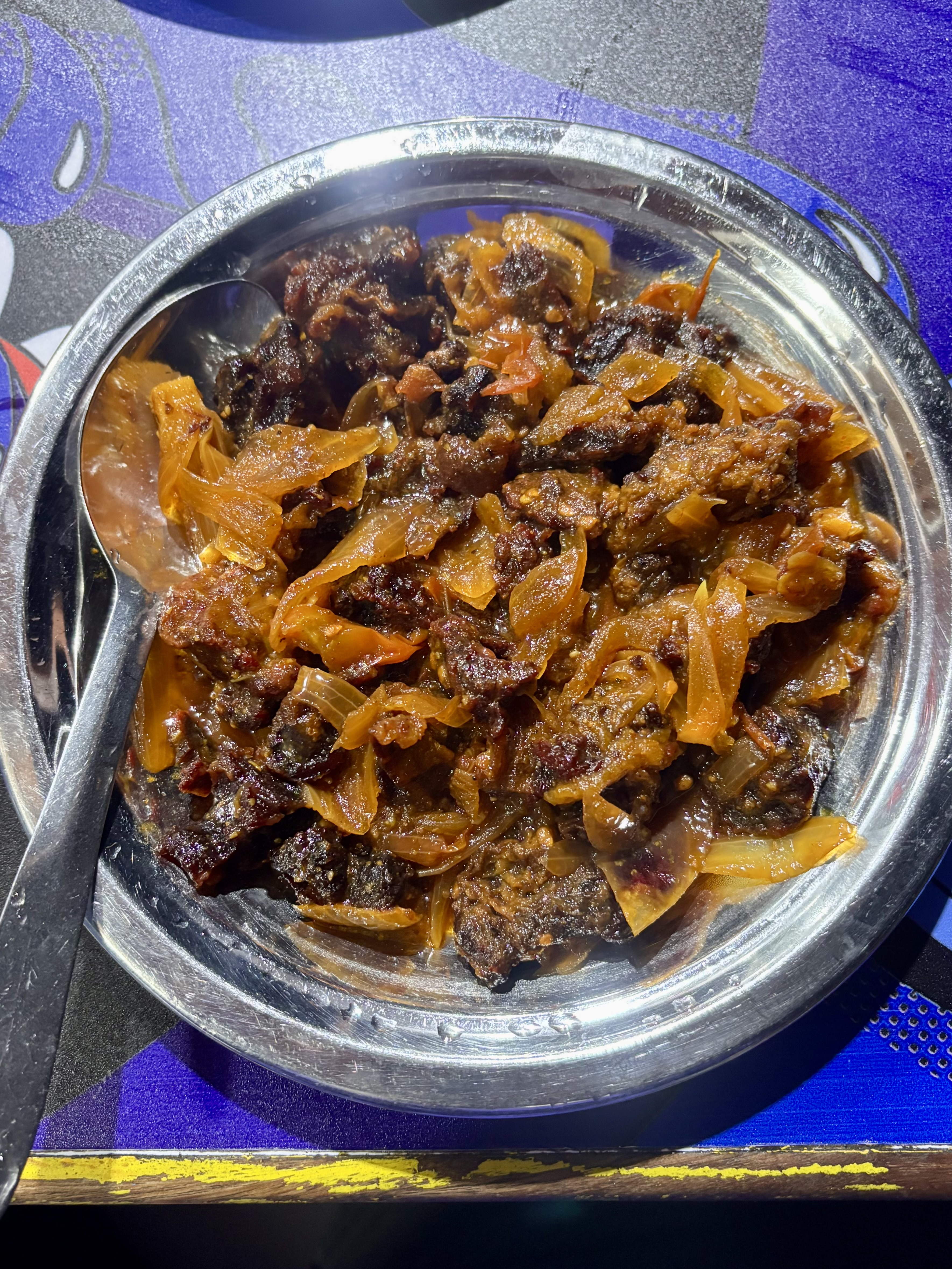 Dark caramelized meat cooked with golden onions in a steel bowl, rich with spices.