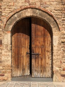 A a half-open wooden door with upper arch surrounded by a stone wall