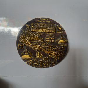 A bronze plaque, probably fridge magnet, displays a detailed map of Nepal, highlighting major mountain ranges like Everest and Annapurna and cultural sites such as Swayambhunath and Pashupatinath against a white background.