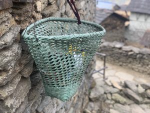 A green woven basket with a mesh pattern hangs on a stone wall, containing small colorful items.