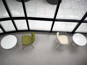 Overhead view of a minimalist setting with two round white tables and modern chairs, one green and one white, next to large glass windows.