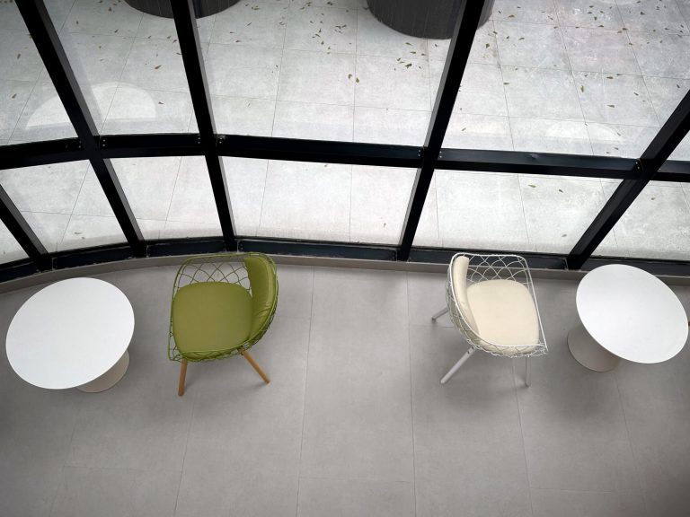 Overhead view of a minimalist setting with two round white tables and modern chairs, one green and one white, next to large glass windows.