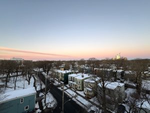A high-angle, wide shot looking over a snow-covered city street and neighborhood. Colorful houses with snow on their roofs run along an asphalt road, surrounded by bare trees. The horizon features a soft pink and orange glow transitioning into a clear blue sky. 