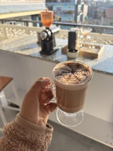 A hand holds a glass mug of frothy cappuccino against a sunny balcony backdrop.
