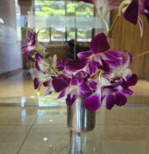 A beautiful bouquet of orchids with delicate purple-and-white petals arranged in a shiny metal vase. Soft natural light highlights the flowers and creates gentle reflections on the glass table, while the blurred greenery in the background adds a calm indoor atmosphere.