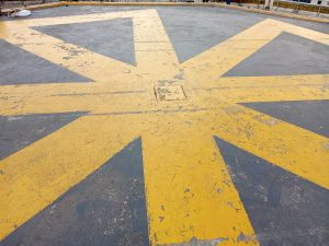 A weathered yellow eight-pointed star design painted on a Helipad concrete surface, featuring a square metal access hatch positioned directly in its center.