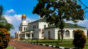 Side profile view of the historic Mengo Palace (Lubiri) in Kampala, showcasing its white colonial-style architecture, prominent golden dome tower, green tiled roofs, and well-manicured green lawns under a bright blue sky.