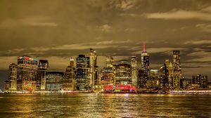 The Lower Manhattan skyline at night as seen across the East River, with glittering skyscrapers reflecting golden light on the water, the Empire State Building lit with a pink spire, and a dramatic overcast sky, New York City, USA. 