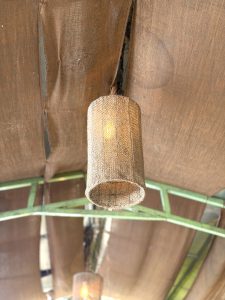 Cylindrical burlap/jute pendant light shade glowing warmly, hanging from a fabric-draped ceiling with green metal framework.
