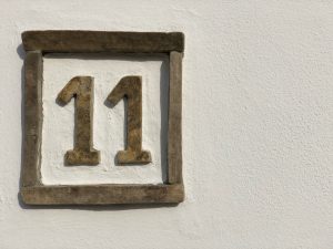 A square house number plaque is mounted on a white textured wall. The plaque has a thick, weathered stone frame in a tan hue. Inside the frame, the number "11" is rendered in large, raised, three-dimensional numerals. These numbers share the same textured stone appearance as the frame, showing signs of age and wear.