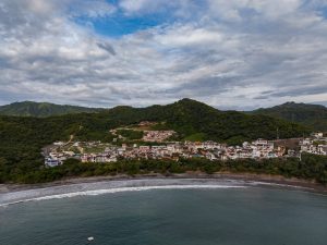 An aerial view of a coastal town between the ocean and green hills. Waves reach the shoreline in the foreground, while colorful buildings sit at the base of forested mountains under a cloudy blue sky.