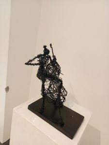 A black wire sculpture of a figure playing a chelo on a pedestal.
