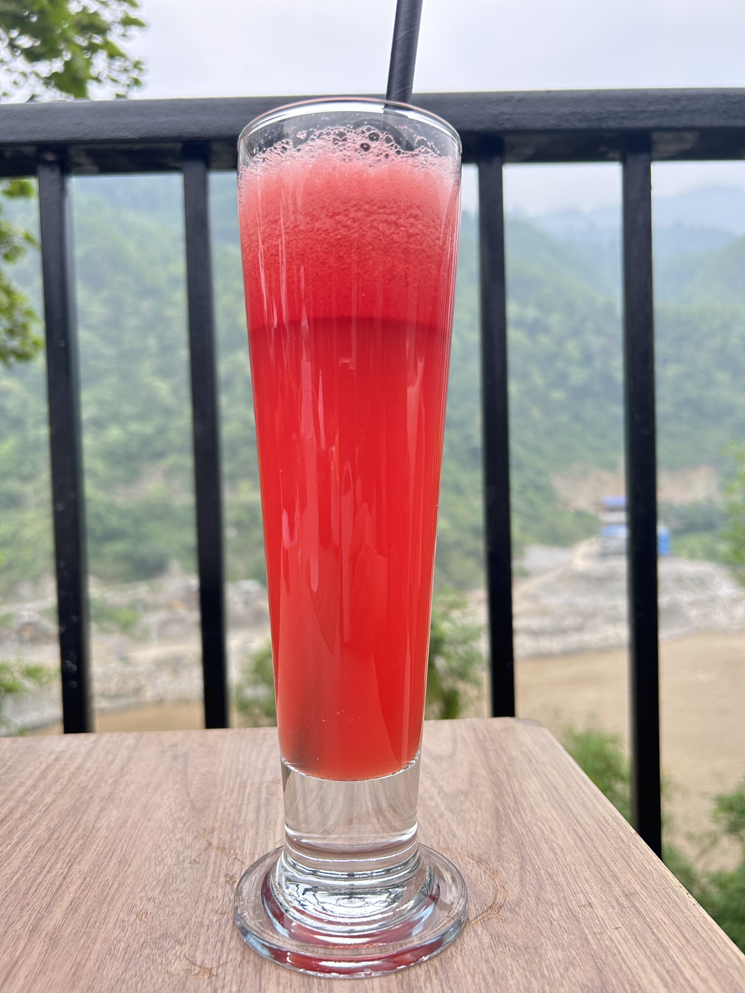 A tall glass of vibrant red watermelon juice with frothy top sits on a wooden table. A black straw stands out against a scenic backdrop of lush green hills.