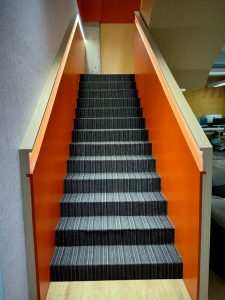 A staircase with gray striped steps and bright orange sides, viewed from the bottom looking up.