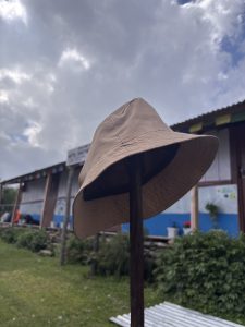 A beige bucket hat placed on a wooden stick in front of a mountain teahouse decorated with colorful prayer flags under a cloudy sky.
