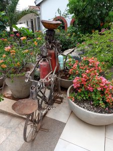 An intricate sculpture of a person riding a bicycle fashioned out of assorted recycled scrap metal materials, featuring old gas cylinders for the torso parts and spark plugs lining the handles, pedals, and seat structures.