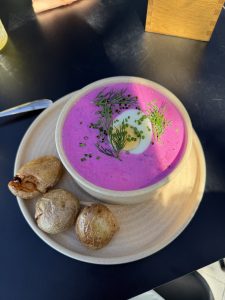 A creamy bowl of vibrant pink soup sits on a sandy-colored, round plate. The soup is garnished with fresh green herbs, including dill and chives, and features a sliced boiled egg in the center. 