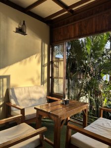 Sun-drenched cafe corner with warm wooden furniture and white cushions, tropical palms spilling through open French doors 