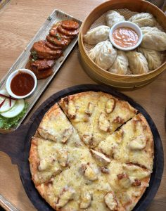 A top-down view of a delicious spread on a wooden table, featuring a golden chicken pizza cut into slices, a round bamboo steamer packed with fresh momos surrounding a dipping sauce, and a long plate of seasoned, sauce-coated potato wedges or cutlets. 
