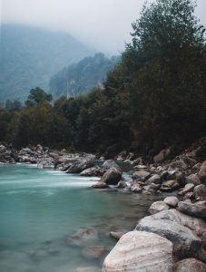 Silky turquoise river winding through smooth boulders and dense forest beneath fog-draped mountain cliffs 