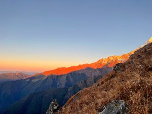 A mountain range glowing vibrant orange at sunrise or sunset under a clear blue sky, viewed from a grassy, rocky hillside in the foreground.