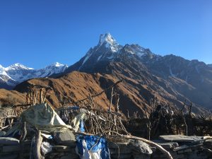 A breathtaking mountain landscape featuring towering, snow-capped peaks against a clear blue sky. In the foreground, a rustic, stone-and-branch structure covered with tattered fabric sits amidst rolling brown hills.