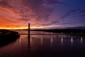 A cable-stayed bridge crosses calm water at sunset, with streetlights reflecting on the surface. The sky is filled with vivid orange, pink, purple, and blue tones, while distant mountains appear as dark silhouettes.
