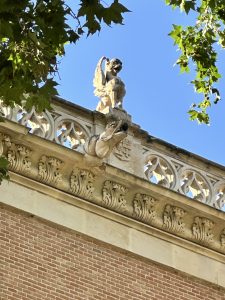Ornate stone gargoyle on a historic building's edge, surrounded by decorative carvings. Bright blue sky and green leaves frame the scene.
