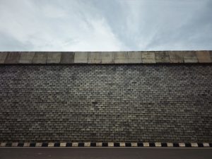 A large stone retaining wall stretches along a roadside beneath a cloudy sky. The repeating block pattern creates an interesting geometric design, giving the image a clean and minimalist look. Captured in Kozhikode, Kerala. 