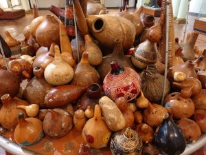 A large collection of dried, hollowed-out gourds and calabashes is clustered together on a circular table. Many gourds feature natural tan and brown tones, while several are decorated with intricate geometric carvings, painted red patterns depicting animals and dark polished finishes.