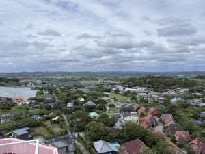 An elevated, wide view of the landscape of Isumi, showing a countryside town with colorful rooftops nestled among lush green trees and fields under a cloudy sky.