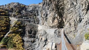 A narrow suspension bridge made of wooden planks and metal cables spans a deep rocky gorge, connecting steep cliffs with a winding stone pathway and wooden railings. Several people walk along the cliffside under a clear blue sky. 