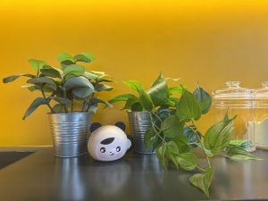 Two artificial plants and a cute panda-shaped object sit on a countertop against a vibrant yellow wall.