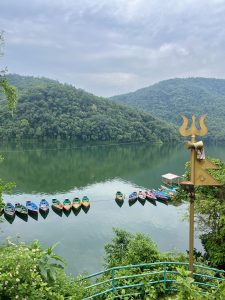 Serene view of Fewa Lake in Pokhara, with colorful boats, green hills, and calm waters beneath a cloudy sky. 