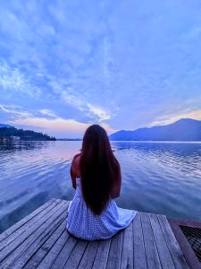 A woman with long hair wearing a blue and white checkered dress sits on a wooden dock, looking out at a peaceful lake surrounded by mountains at dusk.
