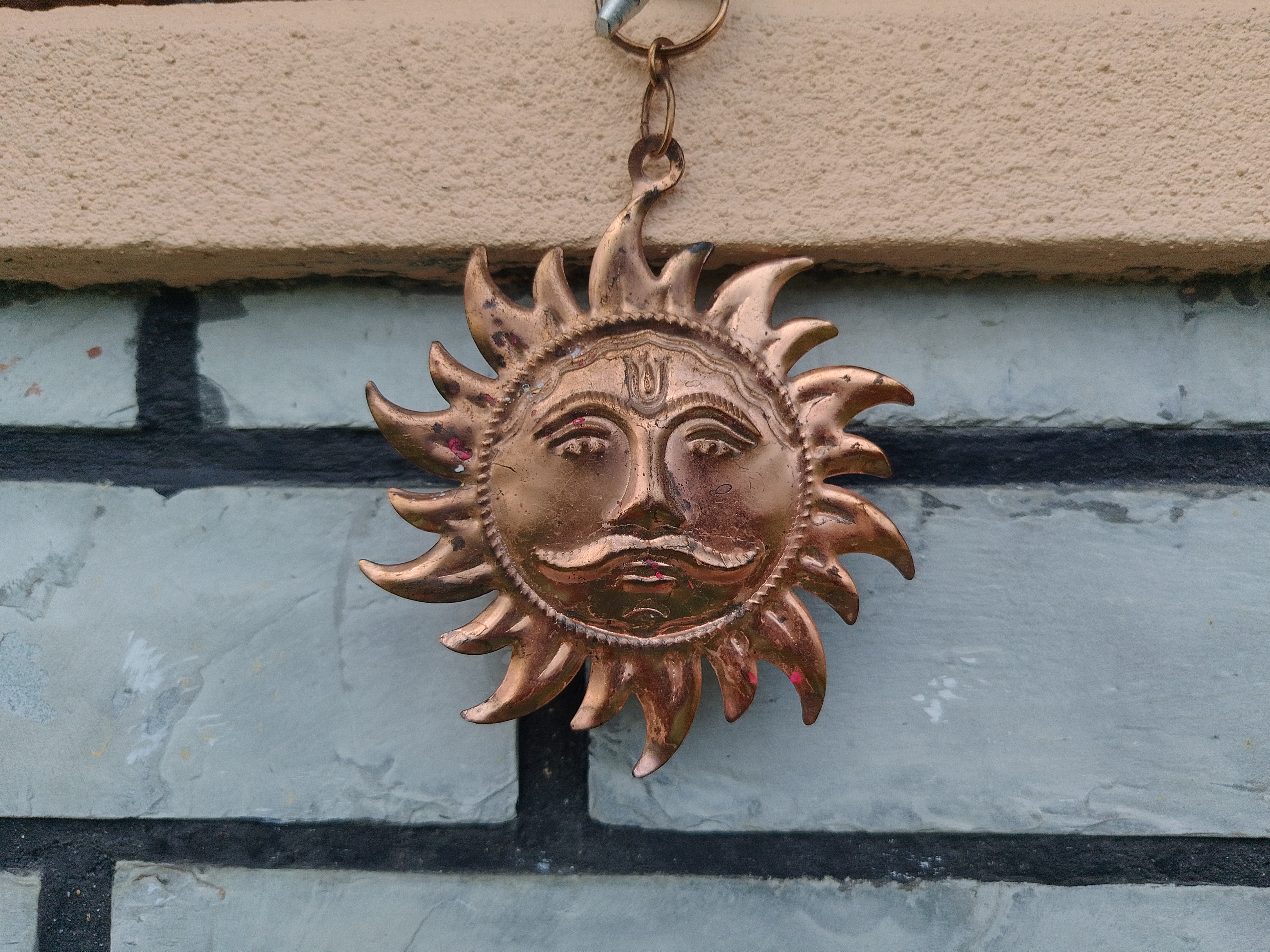 Brass Surya (Sun God) wall medallion with a serene face and flaming rays, hanging from a chain on a stone brick wall.