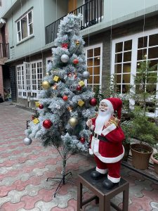 A Santa Claus figure stands on a black stool next to a white-flocked Christmas tree decorated with ornaments. They are located in an outdoor paved courtyard next to a building. 