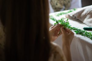 A girl weaving a green plant garland by hand on a white-covered table, with greenery and craft materials arranged nearby.
