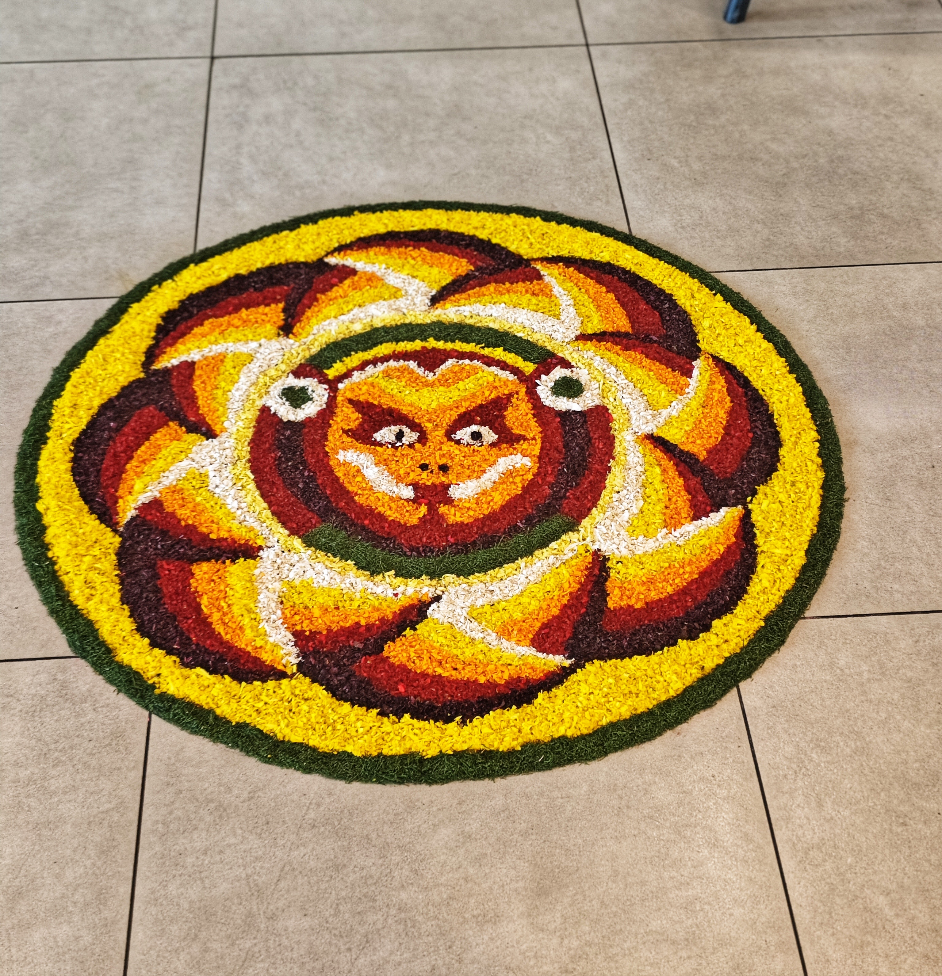 A vibrant circular flower rangoli with a central sun-like face in orange, surrounded by petal patterns in red, yellow, and white on a tiled floor.