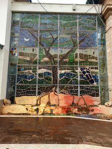 A large public mosaic display composed of multi-colored glass tiles depicting a stylized tree of life, with its canopy set against a blue sky and its root system extending into patterned ground layers. 
